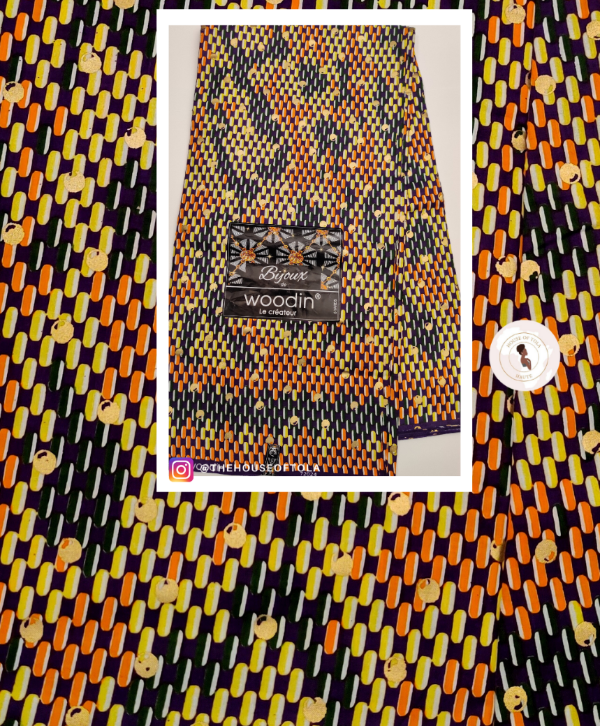 A folded fabric sample with a bold geometric ikat pattern in orange, yellow, black, and white, featuring a 'woodin' brand label. The fabric is presented in a white-bordered frame against a matching patterned background.
